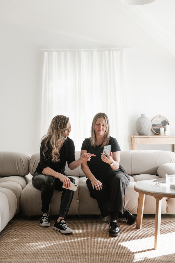 Two blond women sitting on a couch looking at a phone screen and talking.