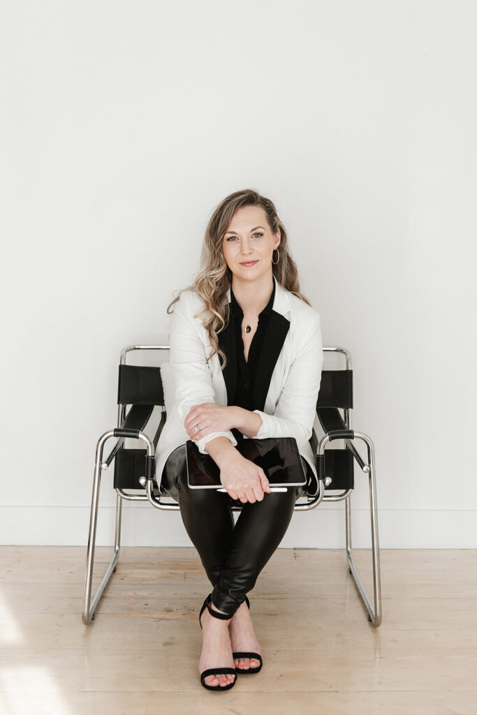 Woman with long blond hair sitting on a black chair against a white wall holding an iPad and smiling