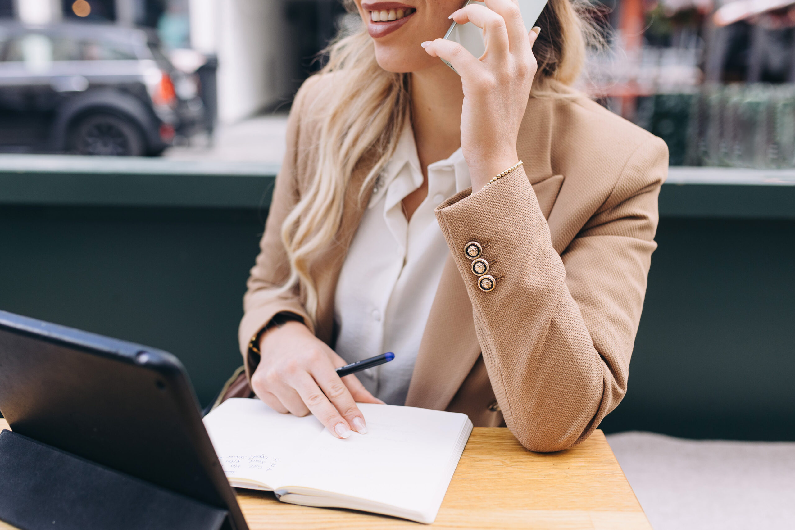 Blonde woman wearing a tan jacket talking on a mobile phone with a notebook, pen, and laptop in front of her.