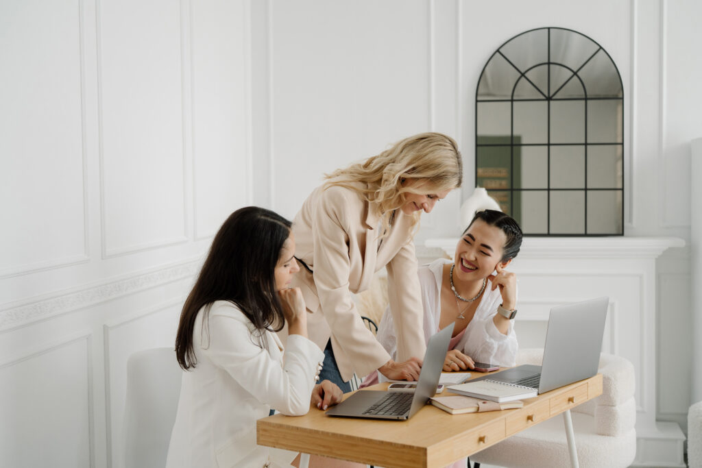 Three women of different ethnicities discussing work at a desk with laptops and notebooks.