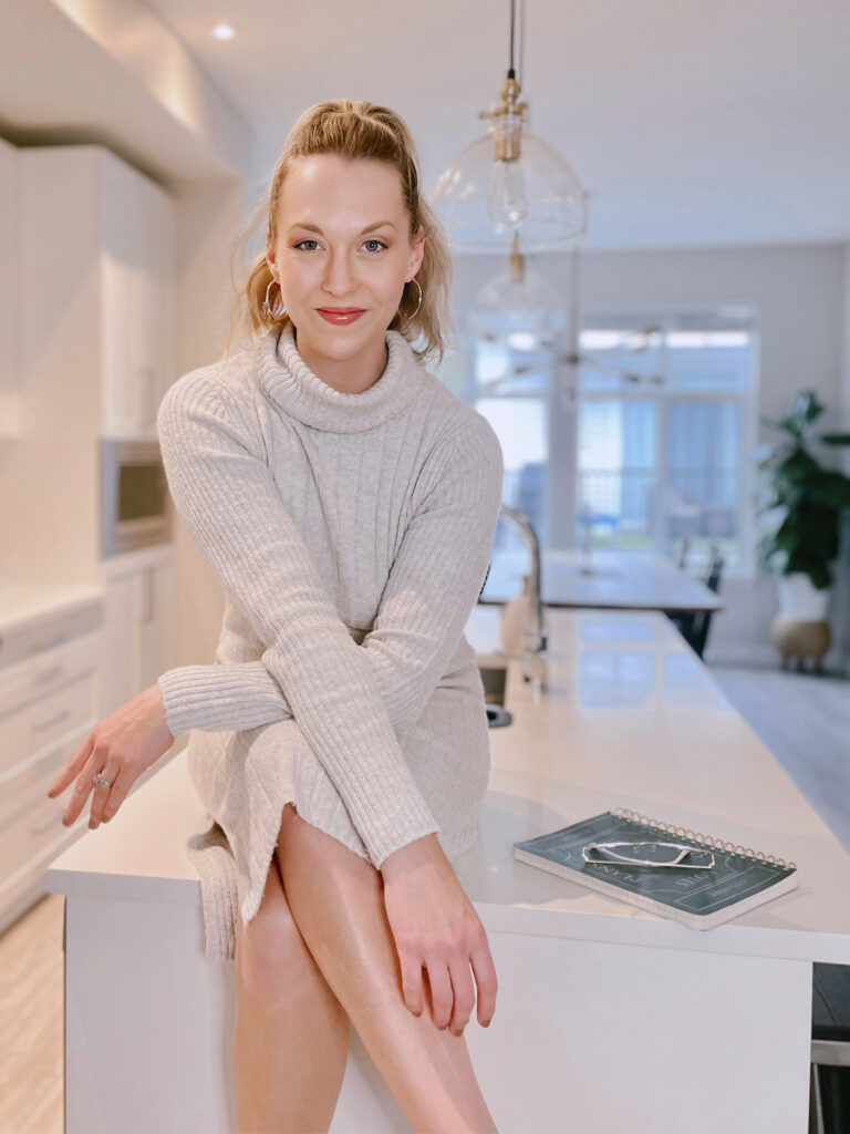 Blonde woman wearing a grey dress sitting on a white countertop smiling.